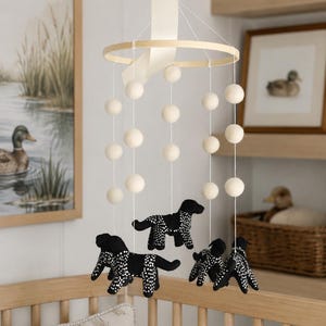 May include: A hanging mobile featuring a circular wooden frame suspended by a cream ribbon. White felt balls are strung on thin white cords, interspersed with four black dog figures decorated with small white polka dots throughout their bodies.