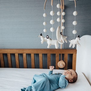 May include: A white cot with a wooden headboard and a white mobile hanging above it. The mobile has white pom-poms and felt animal figures. A baby is lying in the cot looking up at the mobile.