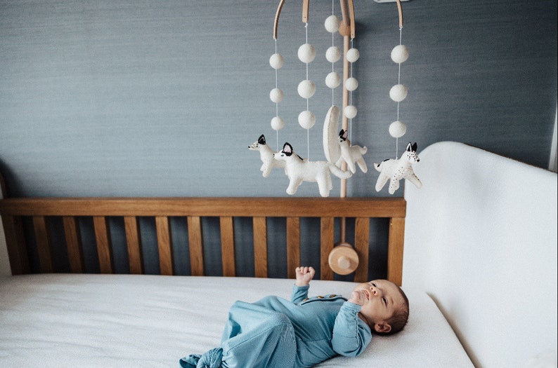 May include: A white cot with a wooden headboard and a white mobile hanging above it. The mobile has white pom-poms and felt animal figures. A baby is lying in the cot looking up at the mobile.