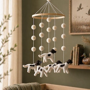 May include: A baby mobile featuring a wooden ring and hanging white felt balls. Below, are four plush dog figures with black spots and brown ears. The mobile hangs above a wooden crib with a green blanket.