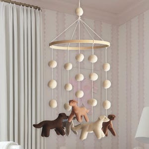K&ouml;nnte beinhalten: Ein h&auml;ngendes Mobile f&uuml;r das Kinderzimmer mit einem runden Holzrahmen, der an wei&szlig;en Schn&uuml;ren befestigt ist. Es besteht aus mehreren cremefarbenen Filzkugeln und vier Filzhunden in Braun-, Beige- und Cremet&ouml;nen, die in unterschiedlichen L&auml;ngen herabh&auml;ngen.