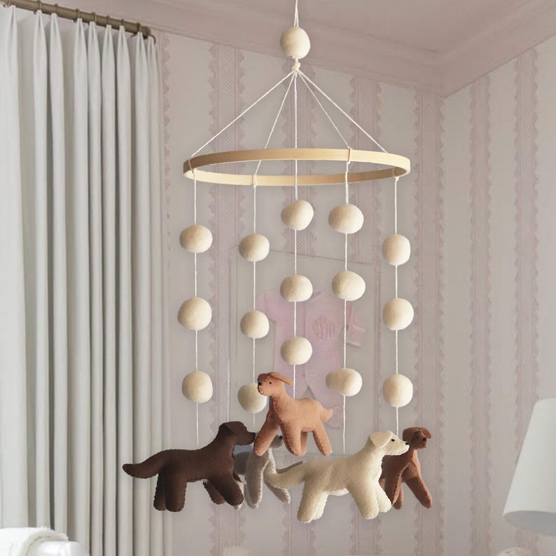K&ouml;nnte beinhalten: Ein h&auml;ngendes Mobile f&uuml;r das Kinderzimmer mit einem runden Holzrahmen, der an wei&szlig;en Schn&uuml;ren befestigt ist. Es besteht aus mehreren cremefarbenen Filzkugeln und vier Filzhunden in Braun-, Beige- und Cremet&ouml;nen, die in unterschiedlichen L&auml;ngen herabh&auml;ngen.