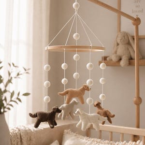 Op de afbeelding: Een houten baby mobile met vilten honden in verschillende kleuren, waaronder bruin, beige en wit. De mobile hangt aan een houten arm en is versierd met witte pompons. De kinderkamerdecoratie omvat een babybedje en een ingelijste print met de tekst "you are so loved."