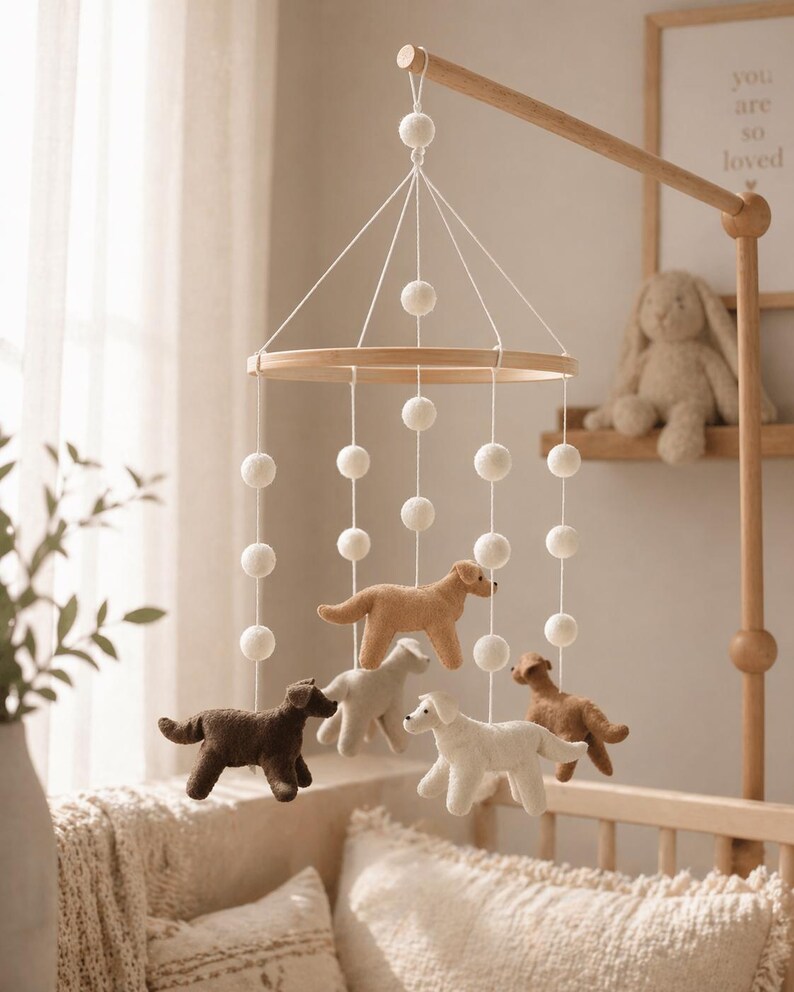 Op de afbeelding: Een houten baby mobile met vilten honden in verschillende kleuren, waaronder bruin, beige en wit. De mobile hangt aan een houten arm en is versierd met witte pompons. De kinderkamerdecoratie omvat een babybedje en een ingelijste print met de tekst "you are so loved."