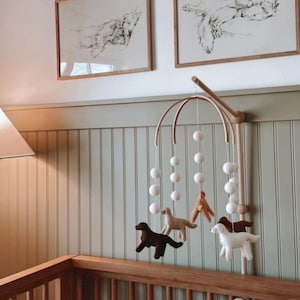 May include: A wooden baby mobile with white felt balls and four felt dog figures in brown, tan, and white hanging from a wooden arch. The mobile is attached to a wooden stand and is hanging above a wooden cot.