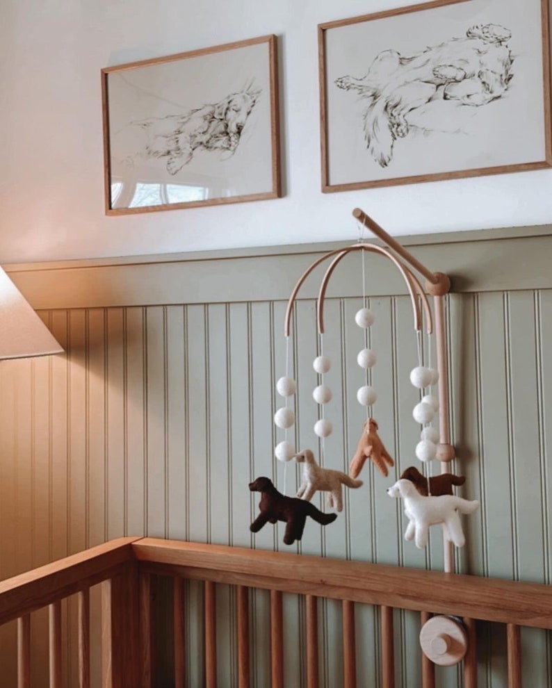 May include: A wooden baby mobile with white felt balls and four felt dog figures in brown, tan, and white hanging from a wooden arch. The mobile is attached to a wooden stand and is hanging above a wooden cot.