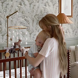 Op de afbeelding: Een kinderkamersc&egrave;ne met een houten wieg, een mobiel met dierenfiguren en een persoon die een baby vasthoudt. De baby draagt een gestreepte outfit en een speen. De persoon draagt een roze en wit gestreepte pyjama.