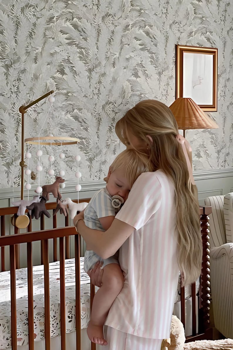 Op de afbeelding: Een kinderkamersc&egrave;ne met een houten wieg, een mobiel met dierenfiguren en een persoon die een baby vasthoudt. De baby draagt een gestreepte outfit en een speen. De persoon draagt een roze en wit gestreepte pyjama.