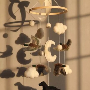 Op de afbeelding: Handgemaakte baby mobile met een houten ring, vilten wolken, manen en kleine vogels. Opgehangen aan witte draad en een cr&egrave;me lint. Een zwart hondenfiguur hangt aan de onderkant.