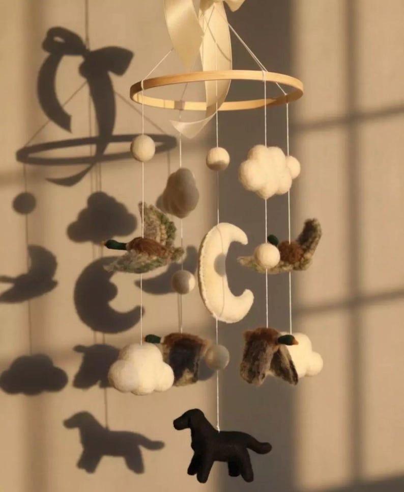 Op de afbeelding: Handgemaakte baby mobile met een houten ring, vilten wolken, manen en kleine vogels. Opgehangen aan witte draad en een cr&egrave;me lint. Een zwart hondenfiguur hangt aan de onderkant.