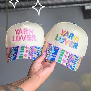 May include: Two off-white baseball caps featuring the embroidered text YARN LOVER. The left cap has pink text, while the right cap has multicolored text. Both caps feature a vibrant, patterned brim with geometric shapes, flowers, and animal prints.