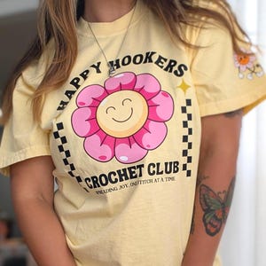 May include: A pale yellow short-sleeve t-shirt featuring a graphic of a pink flower with a smiling face. The text reads HAPPY HOOKERS CROCHET CLUB and SPREADING JOY, ONE STITCH AT A TIME. A black and white checkered border frames the design.