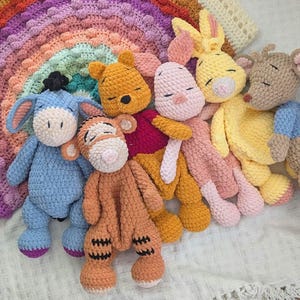 Peut inclure: Une collection de six peluches au crochet représentant des personnages dont un âne bleu, un tigre orange, un ours jaune, un porcelet rose, un lapin jaune et une souris brune, disposés devant un grand arc-en-ciel crocheté coloré.