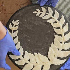 May include: A white silicone mold shaped like a wreath of leaves on a black surface. The mold is being held by gloved hands. The text "Mold Star™ TA-138 Mix by Volume Platin" is visible on a white label.