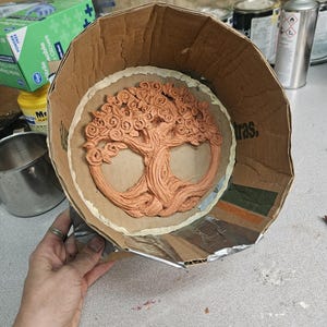 May include: A cardboard mold with a tree of life design made of clay. The mold is lined with aluminum foil and has a brown paper backing.