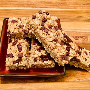May include: A close-up of four granola bars with chocolate chips stacked on a red square plate. The bars are made with oats, nuts, and chocolate chips.