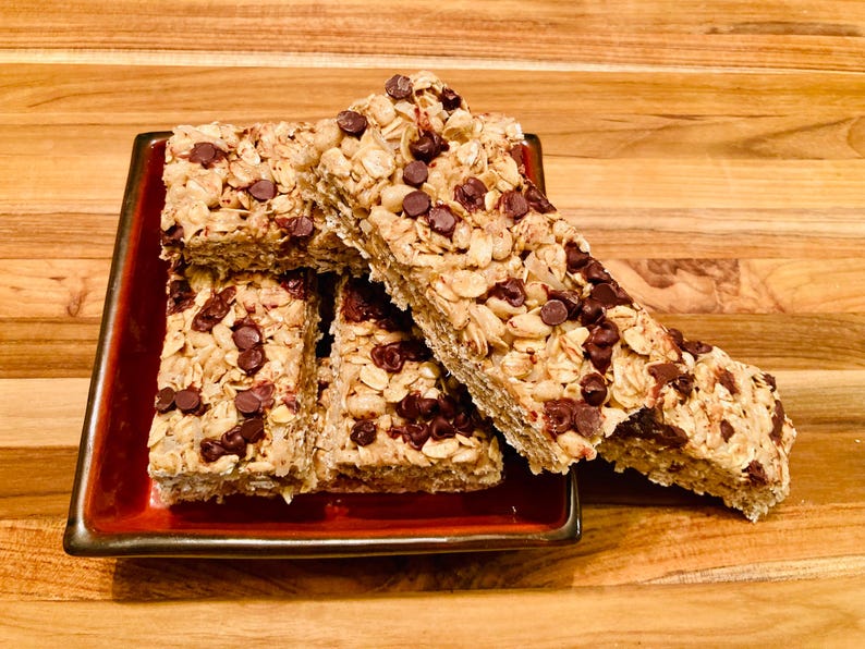May include: A close-up of four granola bars with chocolate chips stacked on a red square plate. The bars are made with oats, nuts, and chocolate chips.