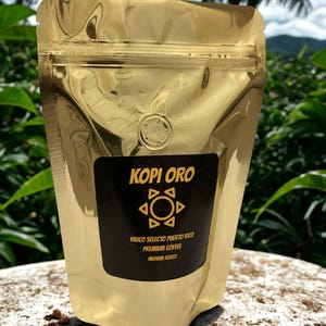 Könnte beinhalten: Ein glänzender goldener Kaffeebeutel mit einem schwarzen Etikett. Das Etikett trägt die Aufschrift "KOPI ORO" mit einem sonnenähnlichen Design und dem Text "Yauco Selecto Puerto Rico Premium Coffee Medium Roast."