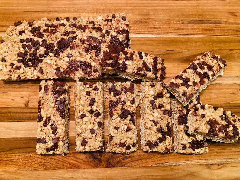 May include: A close-up of a wooden cutting board with several granola bars with chocolate chips. The bars are arranged in a variety of sizes and shapes.