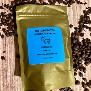 Op de afbeelding: Een gouden foliezak met Haïtiaanse Blue Mountain koffie met het label "Bull Terrier Barista" met een cartoon-hondlogo en de tekst "Luxury in a Cup, Medium Roast, Notes of Plum, Cinnamon, Sweet Herbs, Net Weight 2 oz (57 g)"