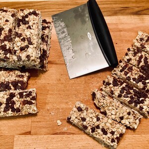 May include: A close-up of a wooden cutting board with a variety of homemade granola bars. The bars are made with oats, nuts, and chocolate chips. A metal spatula is also visible in the image.
