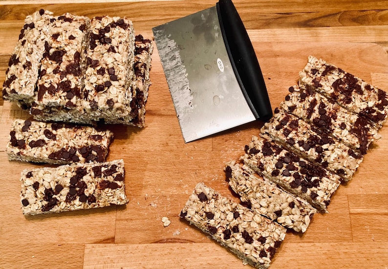 May include: A close-up of a wooden cutting board with a variety of homemade granola bars. The bars are made with oats, nuts, and chocolate chips. A metal spatula is also visible in the image.