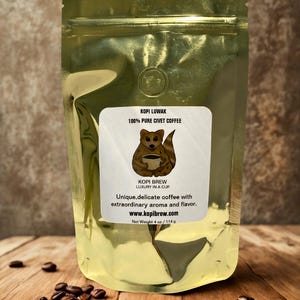 May include: A gold foil coffee bag with a label featuring a civet cat illustration holding a coffee cup. The label reads "KOPI LUWAK 100% PURE CIVET COFFEE" and "KOPI BREW LUXURY IN A CUP". The bag contains 114 g of coffee.