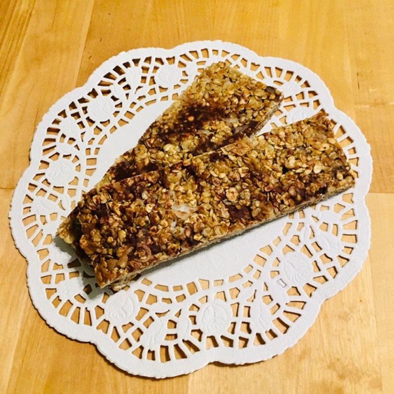 May include: Two granola bars on a white doily. The bars are made with oats, nuts, and seeds. They are a healthy and delicious snack.