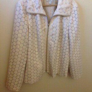 Puede incluir: Chaqueta calada de color crema con cremallera y cuello con solapas. La chaqueta presenta un patrón circular repetitivo. Cuelga de una percha de madera.