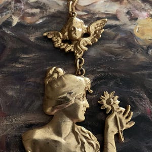 K&ouml;nnte beinhalten: Ein goldfarbener Metallanh&auml;nger mit einem Relief einer Person mit langem Haar, die eine Blume h&auml;lt, gekr&ouml;nt von einer kleinen gefl&uuml;gelten Cherub-Figur. Der Anh&auml;nger h&auml;ngt an einer d&uuml;nnen goldfarbenen Kette und zeigt detaillierte Skulpturen.