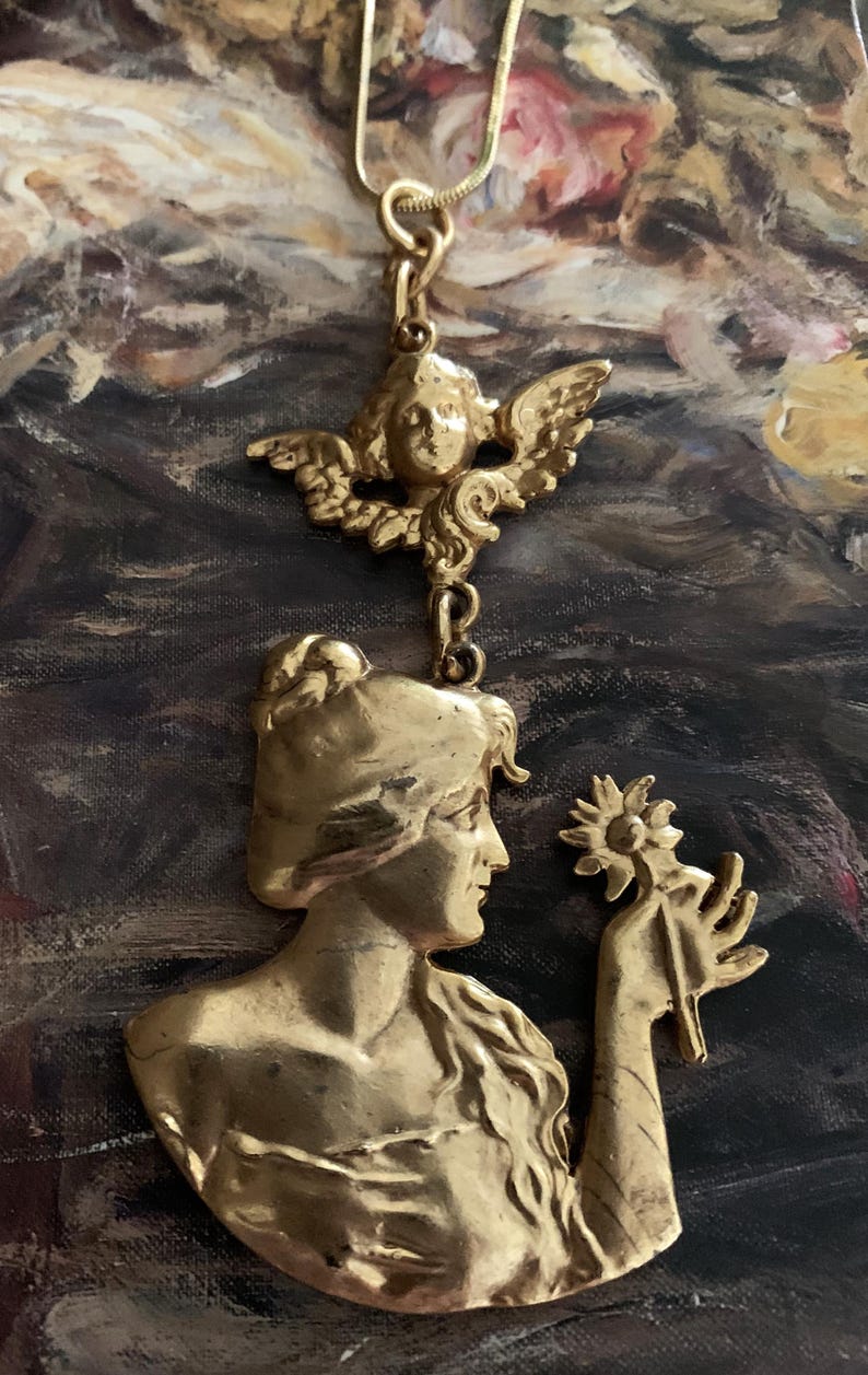 K&ouml;nnte beinhalten: Ein goldfarbener Metallanh&auml;nger mit einem Relief einer Person mit langem Haar, die eine Blume h&auml;lt, gekr&ouml;nt von einer kleinen gefl&uuml;gelten Cherub-Figur. Der Anh&auml;nger h&auml;ngt an einer d&uuml;nnen goldfarbenen Kette und zeigt detaillierte Skulpturen.