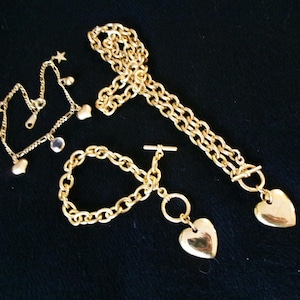 May include: A collection of gold-toned jewelry including a delicate chain bracelet with star and heart charms, a thicker chain bracelet with a toggle clasp and a heart pendant, and a long chain necklace featuring a large heart-shaped pendant.
