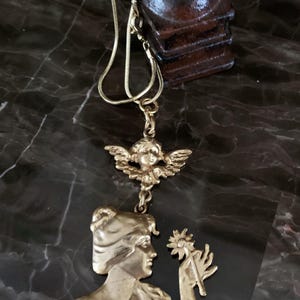 K&ouml;nnte beinhalten: Eine goldfarbene Metallkette mit einem Anh&auml;nger, der das Profil einer Person mit einer Blume zeigt, verbunden mit einem kleinen Cherub-Anh&auml;nger. Der Anh&auml;nger h&auml;ngt an einer d&uuml;nnen, flexiblen goldfarbenen Kette und zeigt ein detailliertes Relief.