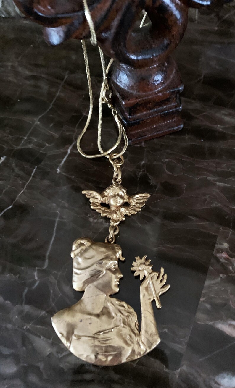 K&ouml;nnte beinhalten: Eine goldfarbene Metallkette mit einem Anh&auml;nger, der das Profil einer Person mit einer Blume zeigt, verbunden mit einem kleinen Cherub-Anh&auml;nger. Der Anh&auml;nger h&auml;ngt an einer d&uuml;nnen, flexiblen goldfarbenen Kette und zeigt ein detailliertes Relief.