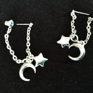 May include: A pair of silver-toned earrings featuring a chain design that connects a post back to a dangling charm set. Each earring includes a small crescent moon charm and a five-pointed star charm suspended from the silver-colored metal chain links.