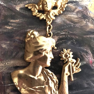 K&ouml;nnte beinhalten: Ein goldfarbener Metallanh&auml;nger mit einem Relief einer Person im Profil, die eine Blume h&auml;lt, verbunden durch einen Biegering mit einem separaten goldfarbenen Metall-Cherub-Ornament mit Fl&uuml;geln, beide mit polierter Oberfl&auml;che.