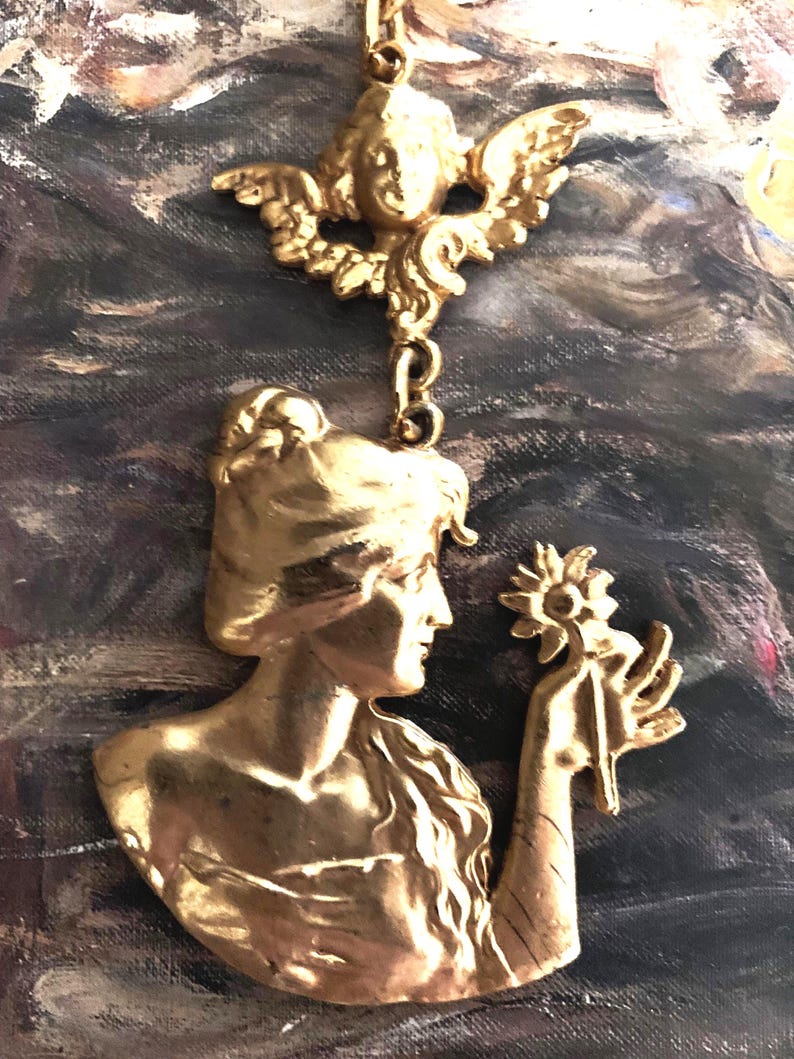 K&ouml;nnte beinhalten: Ein goldfarbener Metallanh&auml;nger mit einem Relief einer Person im Profil, die eine Blume h&auml;lt, verbunden durch einen Biegering mit einem separaten goldfarbenen Metall-Cherub-Ornament mit Fl&uuml;geln, beide mit polierter Oberfl&auml;che.