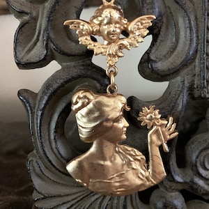 K&ouml;nnte beinhalten: Ein goldfarbener Metallanh&auml;nger mit einem skulptierten Cherub an der Oberseite, der durch einen kleinen Ring mit einem gr&ouml;&szlig;eren unteren Teil verbunden ist, welcher das Profil einer Person mit Hochsteckfrisur und einer Blume zeigt.