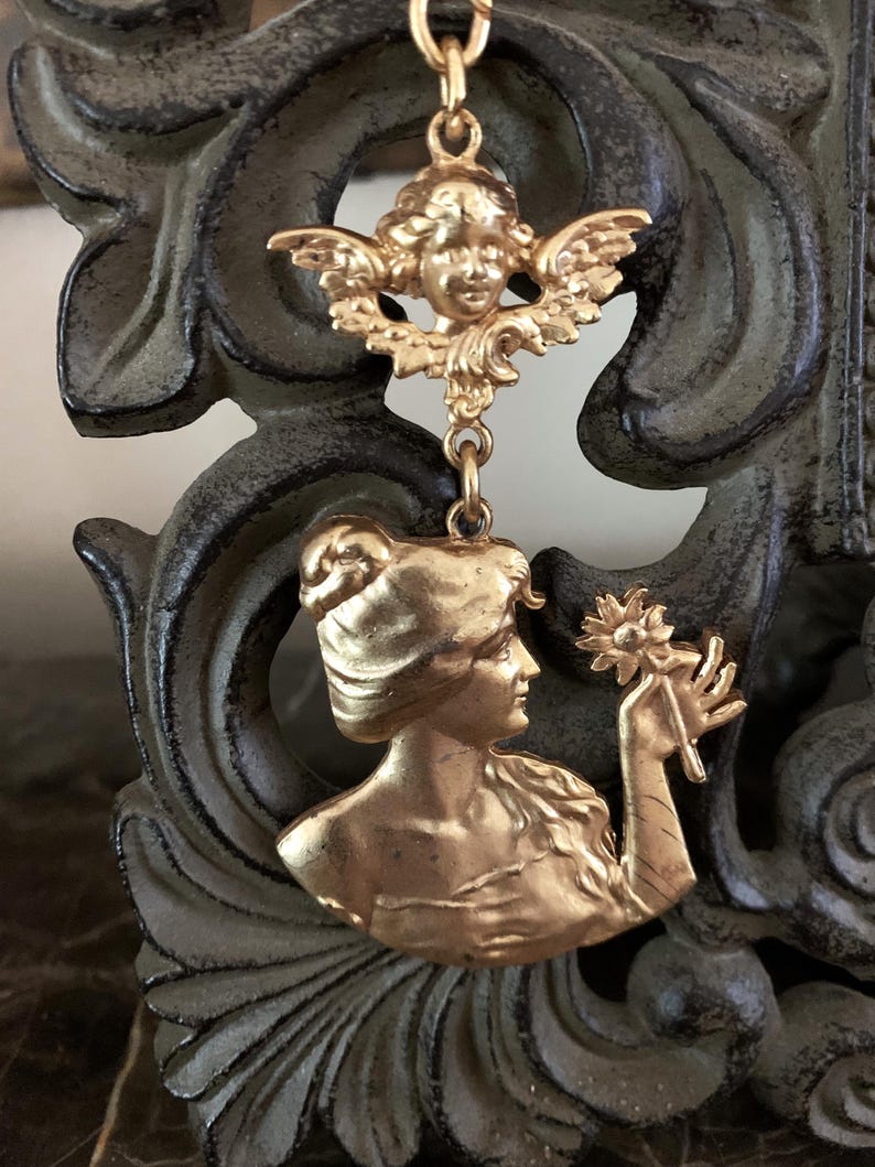 K&ouml;nnte beinhalten: Ein goldfarbener Metallanh&auml;nger mit einem skulptierten Cherub an der Oberseite, der durch einen kleinen Ring mit einem gr&ouml;&szlig;eren unteren Teil verbunden ist, welcher das Profil einer Person mit Hochsteckfrisur und einer Blume zeigt.