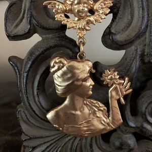K&ouml;nnte beinhalten: Ein goldfarbener Metallanh&auml;nger mit einem Cherub an der Oberseite, der durch einen Verbindungsring mit einem unteren Teil verbunden ist, welcher das Profil einer Person mit Hochsteckfrisur zeigt, die eine einzelne Blume h&auml;lt. Das Metall ist matt.