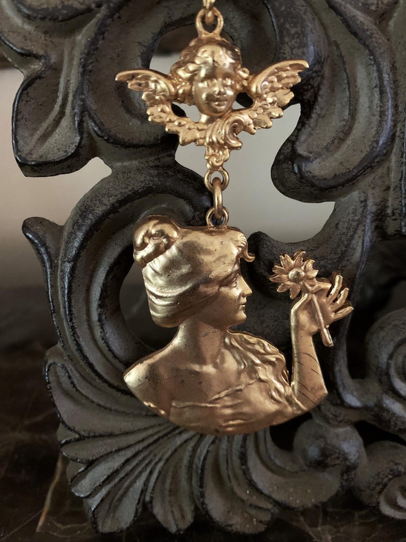 K&ouml;nnte beinhalten: Ein goldfarbener Metallanh&auml;nger mit einem Cherub an der Oberseite, der durch einen Verbindungsring mit einem unteren Teil verbunden ist, welcher das Profil einer Person mit Hochsteckfrisur zeigt, die eine einzelne Blume h&auml;lt. Das Metall ist matt.