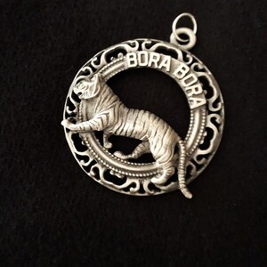 Op de afbeelding: Zilverkleurige hanger met een gedetailleerde tijger in een cirkelvormige lijst. Het woord "BORA BORA" is rond de tijger gegraveerd. De hanger heeft een sierlijk, opengewerkt ontwerp en een kleine lus voor een ketting.