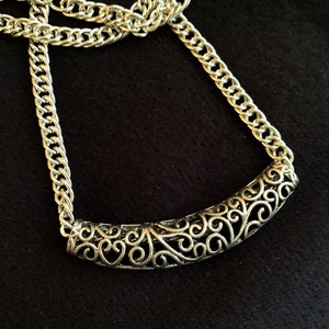 Op de afbeelding: Een zilveren ketting met een delicate, sierlijke hanger. De hanger is een gebogen, opengewerkt ontwerp met een bloemenmotief. De ketting is een eenvoudige, platte schakelstijl.