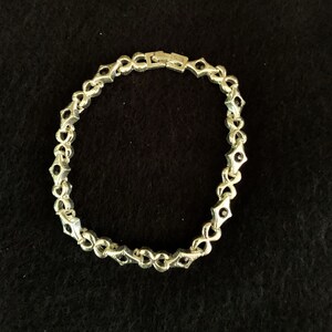 May include: Silver-toned bracelet with a repeating pattern of diamond-shaped links and small black accents. The bracelet is closed with a clasp. The jewelry is displayed on a black surface.
