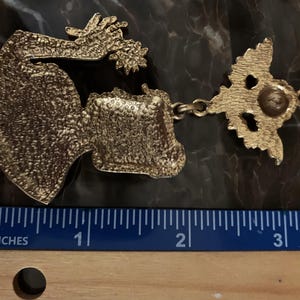 K&ouml;nnte beinhalten: Ein goldfarbener Metallanh&auml;nger mit einer strukturierten, stilisierten Figur mit ausgestrecktem Arm und sternf&ouml;rmigem Element, verbunden mit einer dekorativen &Ouml;se. Ein Lineal darunter zeigt, dass der Anh&auml;nger etwa 8,9 cm lang ist.