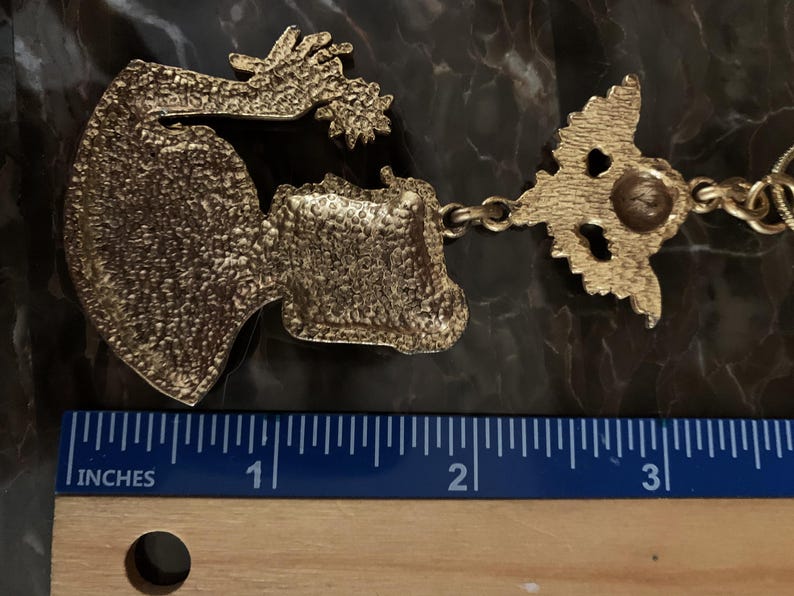K&ouml;nnte beinhalten: Ein goldfarbener Metallanh&auml;nger mit einer strukturierten, stilisierten Figur mit ausgestrecktem Arm und sternf&ouml;rmigem Element, verbunden mit einer dekorativen &Ouml;se. Ein Lineal darunter zeigt, dass der Anh&auml;nger etwa 8,9 cm lang ist.