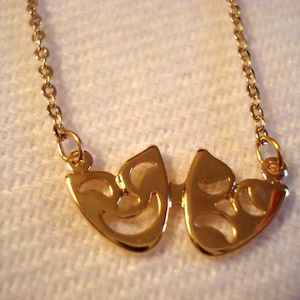 Puede incluir: Un collar dorado con dos máscaras de teatro estilizadas, una sonriendo y otra frunciendo el ceño. Las máscaras están conectadas y suspendidas de una delicada cadena. El collar se muestra sobre una superficie blanca texturizada.