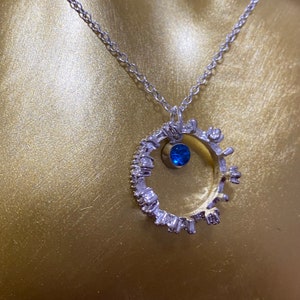 Op de afbeelding: Een zilveren ketting met een ronde hanger met een blauwe edelsteen en een rij kleine heldere stenen rond de rand.