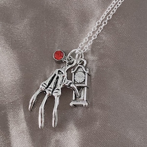 May include: Silver necklace with a red crystal charm, a skeletal hand charm, and a clock charm. The clock charm has a Roman numeral face.