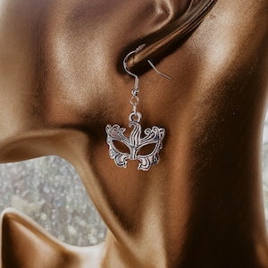 May include: Silver earring with a detailed mask design. The mask is ornate and has a delicate, intricate design.
